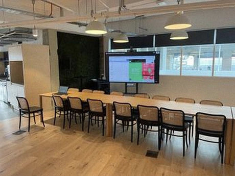 Coinbase London office meeting space with a long shared table, wooden chairs and a large 86-inch LG display on a stand, served by a QSC Core 110f and Shure MXW wireless microphone system, in London, UK.