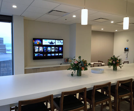 Café at Russell Reynolds Associates' San Francisco office, with a wall-mounted display and soundbar showing the Apple TV home screen above a credenza and a long bar-height table in the foreground.
