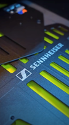Close-up of the front panel of a dark rack-mount Sennheiser wireless unit, photographed at an angle, with the Sennheiser logo and wordmark beside rows of illuminated yellow-green status indicators.