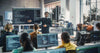 A teacher instructs students in a tech classroom with computers and screens.
